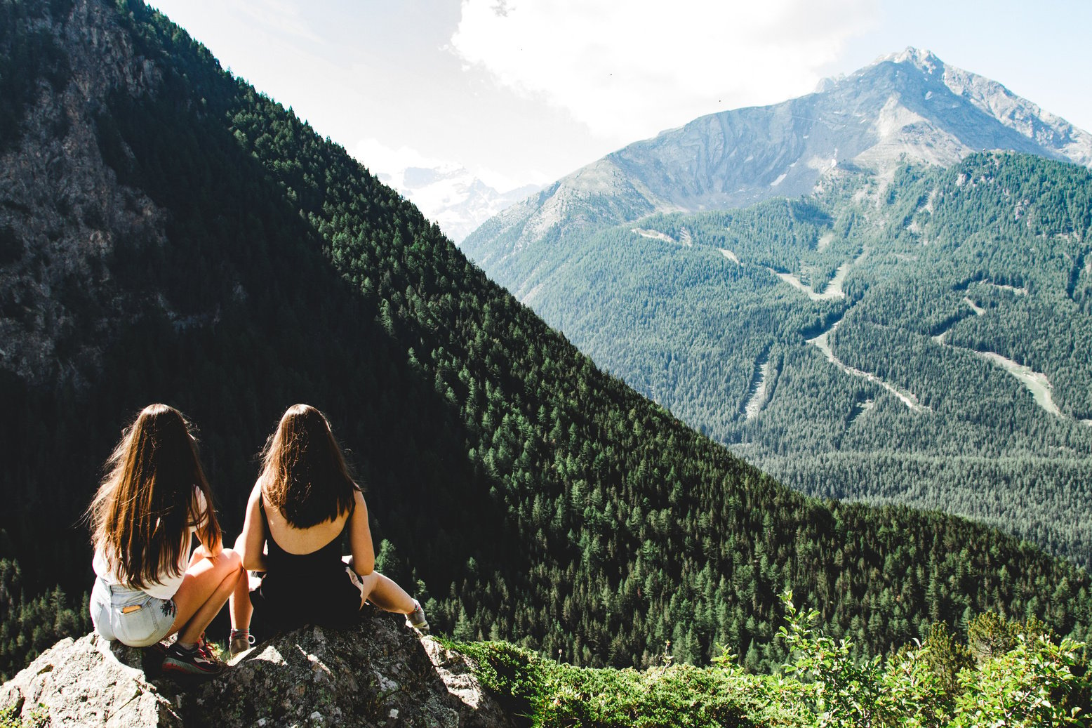 Two Women Sitting on Rock Facing Mountains Under White Cloudy Skies